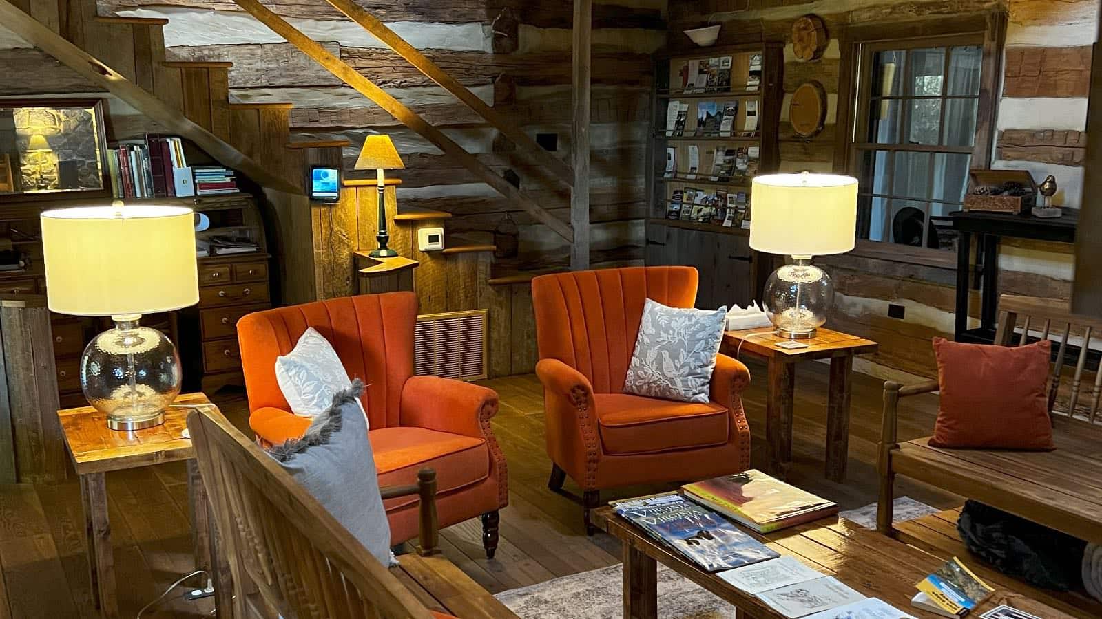 Great room with log cabin walls, hardwood flooring, wooden staircase to second floor, bright orange upholstered armchairs, and wooden benches