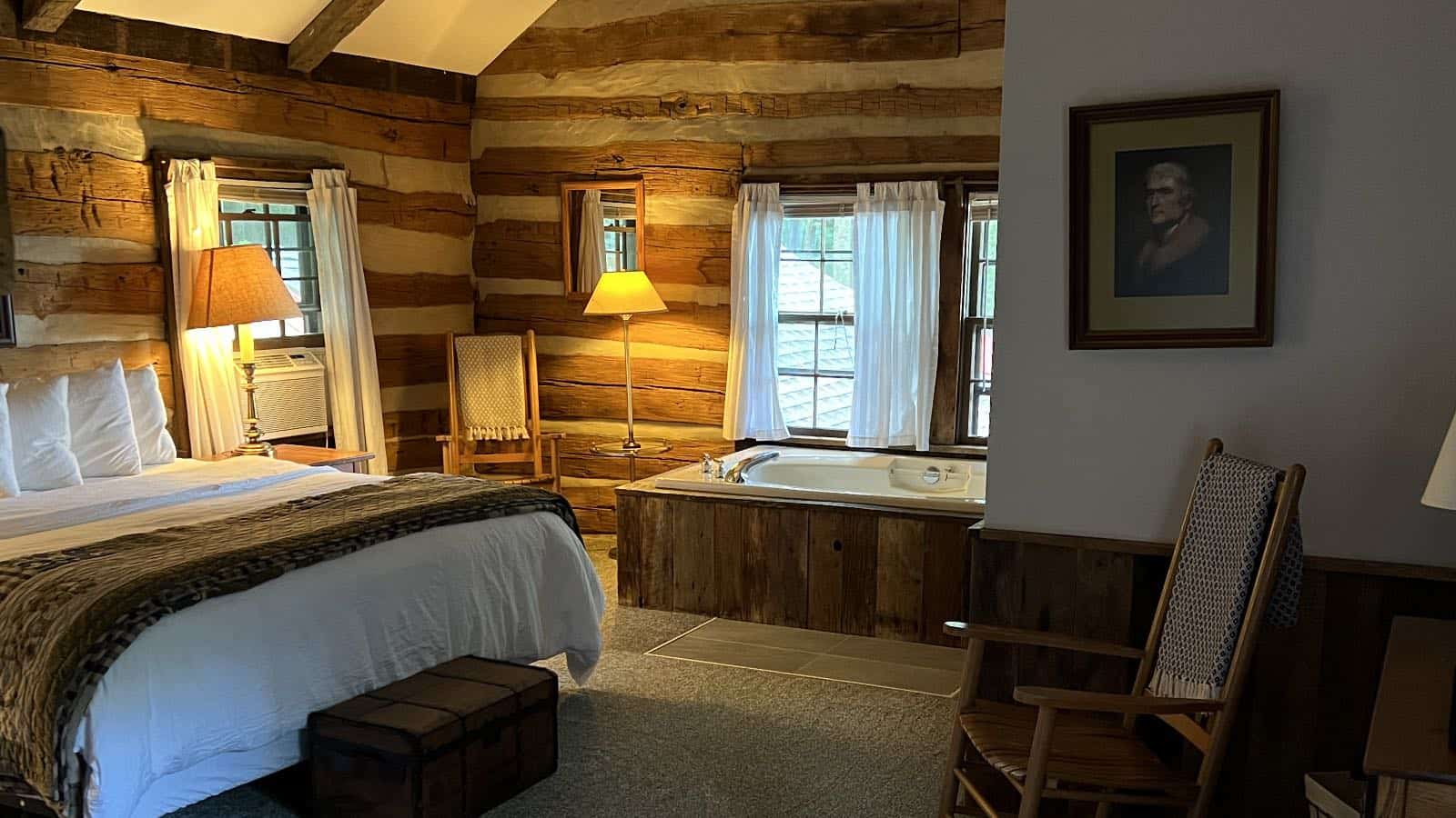 Bedroom with log cabin walls, carpeting, bed with white bedding, wooden rocking chair, and whirlpool tub